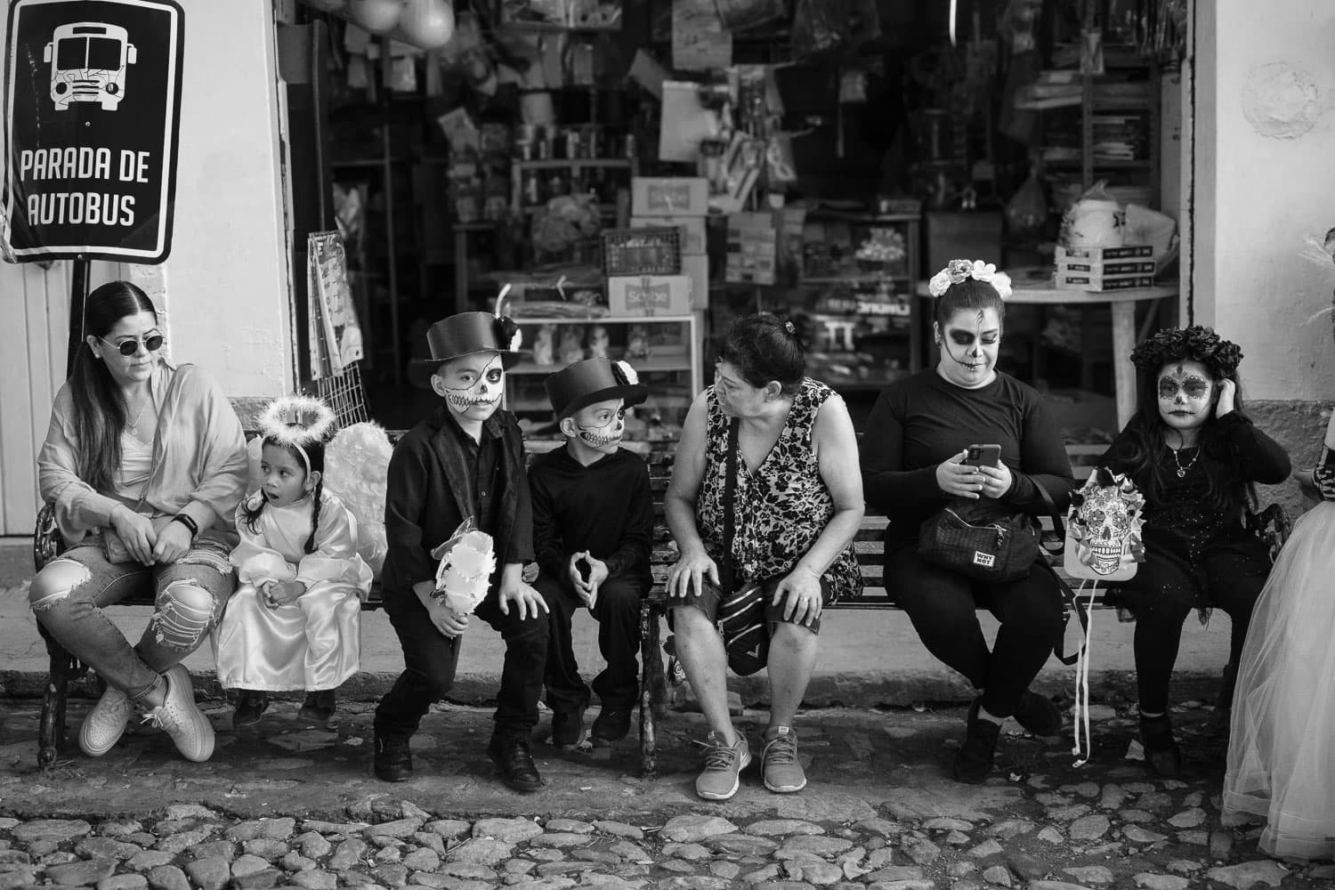 Familes sit at a bus stop while waiting for the Day of the Little Angels parade to start in Ajijic.